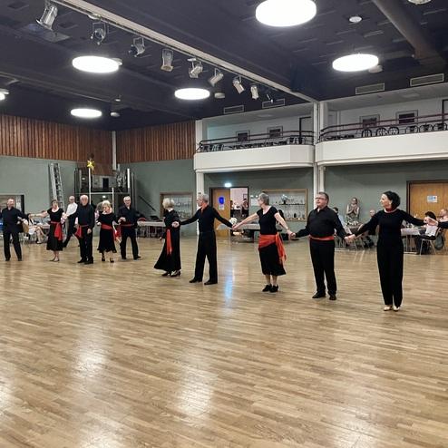 Tanzende Gruppe von Männern und Frauen in schwarzer Kleidung mit roten Bändern auf einer Holzfläche in einem Saal.