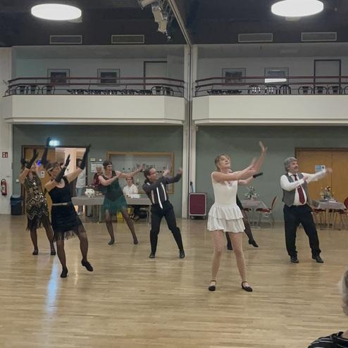 Tanzgruppe in eleganter Kleidung führt eine Choreografie auf einer Tanzfläche vor einem Publikum auf.