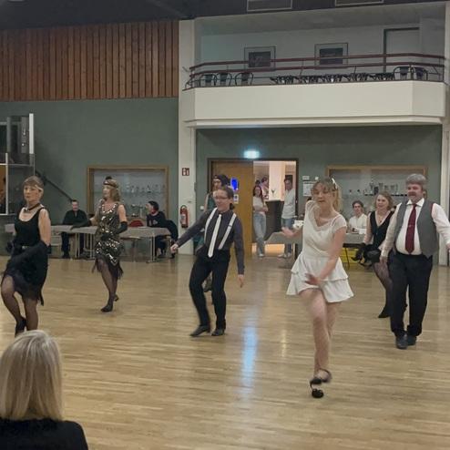 Tanzgruppe in eleganten Kleidern bewegt sich auf einem Holzfußboden während einer Veranstaltung in einem Saal.