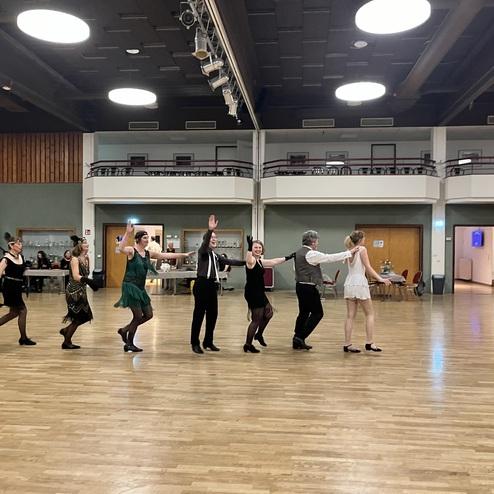 Tanzgruppe in festlicher Kleidung führt eine Choreografie auf einer holzlatten Tanzfläche aus.