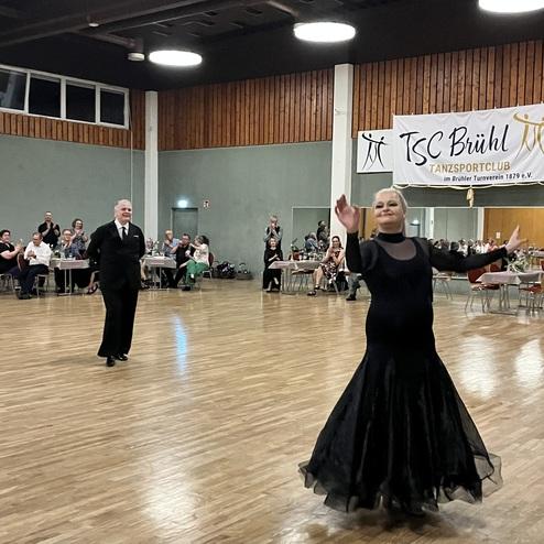 Tanzpaar in eleganter Kleidung führt einen Tanz in einer festlichen Atmosphäre mit Zuschauern im Hintergrund auf.