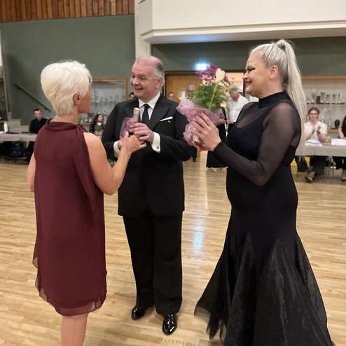 Drei Personen stehen in festlicher Kleidung, eine überreicht Blumen, im Hintergrund ist eine tanzende Menge sichtbar.