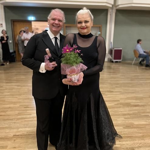 Paar in eleganter Kleidung posiert lächelnd, der Mann hält eine Flasche, die Frau einen Blumenstrauß in Geschenkpapier.