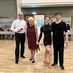 Vier Personen stehen in einem Tanzsaal, lächelnd, mit Pokalen in den Händen und fröhlicher Atmosphäre im Hintergrund.