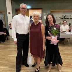 Drei Personen stehen zusammen in einem Raum, eine hält einen Blumenstrauß, Hintergrund zeigt Trophäen und eine weitere Person.