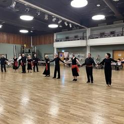 Tanzende Gruppe von Männern und Frauen in schwarzer Kleidung mit roten Bändern auf einer Holzfläche in einem Saal.