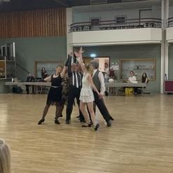 Tanzgruppe in eleganter Kleidung, die auf einer Holzfläche eine tänzerische Aufführung präsentiert.