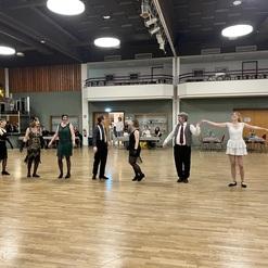 Tanzgruppe in eleganter Kleidung führt eine Choreografie in einem großen Saal mit Holzboden auf.