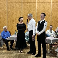 Paar steht auf einer Veranstaltung, während ein Mann mit Mikrofon spricht. Gäste sitzen an Tischen im Hintergrund.