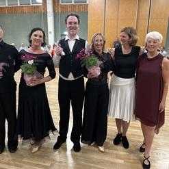 Fünf Personen in eleganter Kleidung posieren lächelnd, halten Blumensträuße und eine Flasche Sekt in einem festlichen Saal.