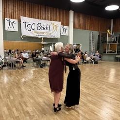 Zwei Frauen umarmen sich in einem Festsaal des Tanzsportclubs Brühl, im Hintergrund tanzende Gäste und Dekoration.