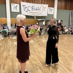 Zwei Frauen stehen sich lächelnd gegenüber, eine überreicht der anderen einen Blumenstrauß, im Hintergrund ein festlich dekorierter Raum.