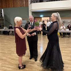 Zwei Frauen überreichen ein Geschenk mit Blumen, während ein Mann im Hintergrund lächelt. Tanzveranstaltung in einem Saal.