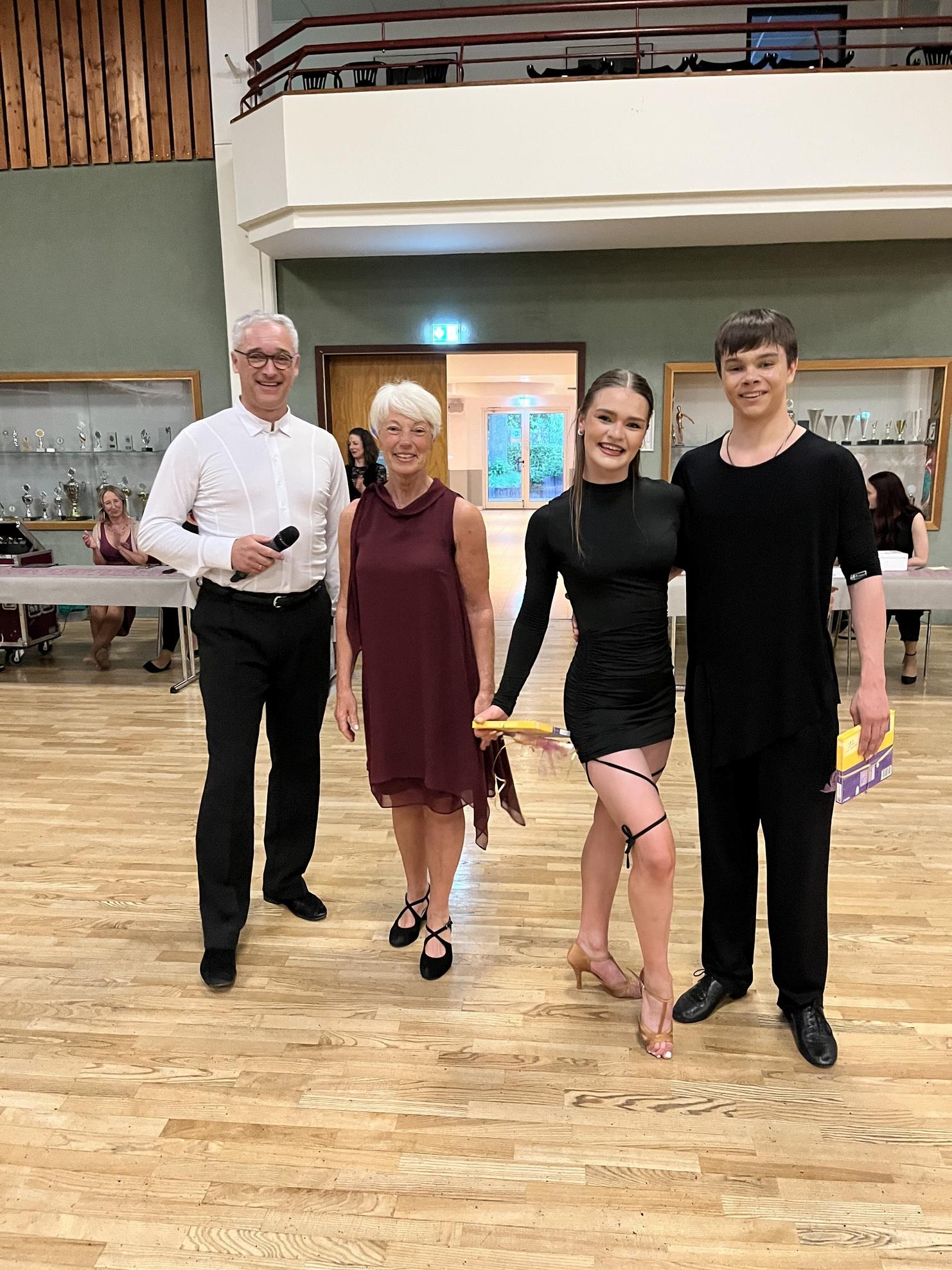 Vier Personen stehen in einem Tanzsaal, lächelnd, mit Pokalen in den Händen und fröhlicher Atmosphäre im Hintergrund.