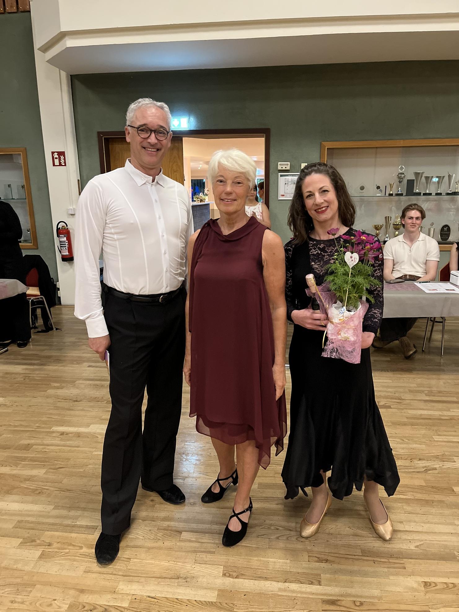Drei Personen stehen zusammen in einem Raum, eine hält einen Blumenstrauß, Hintergrund zeigt Trophäen und eine weitere Person.