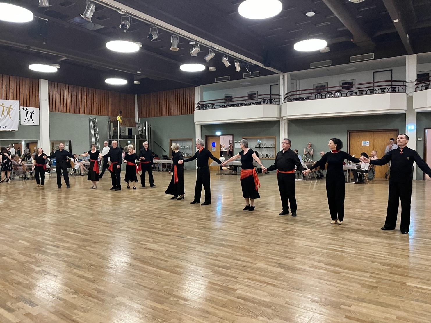 Tanzende Gruppe von Männern und Frauen in schwarzer Kleidung mit roten Bändern auf einer Holzfläche in einem Saal.