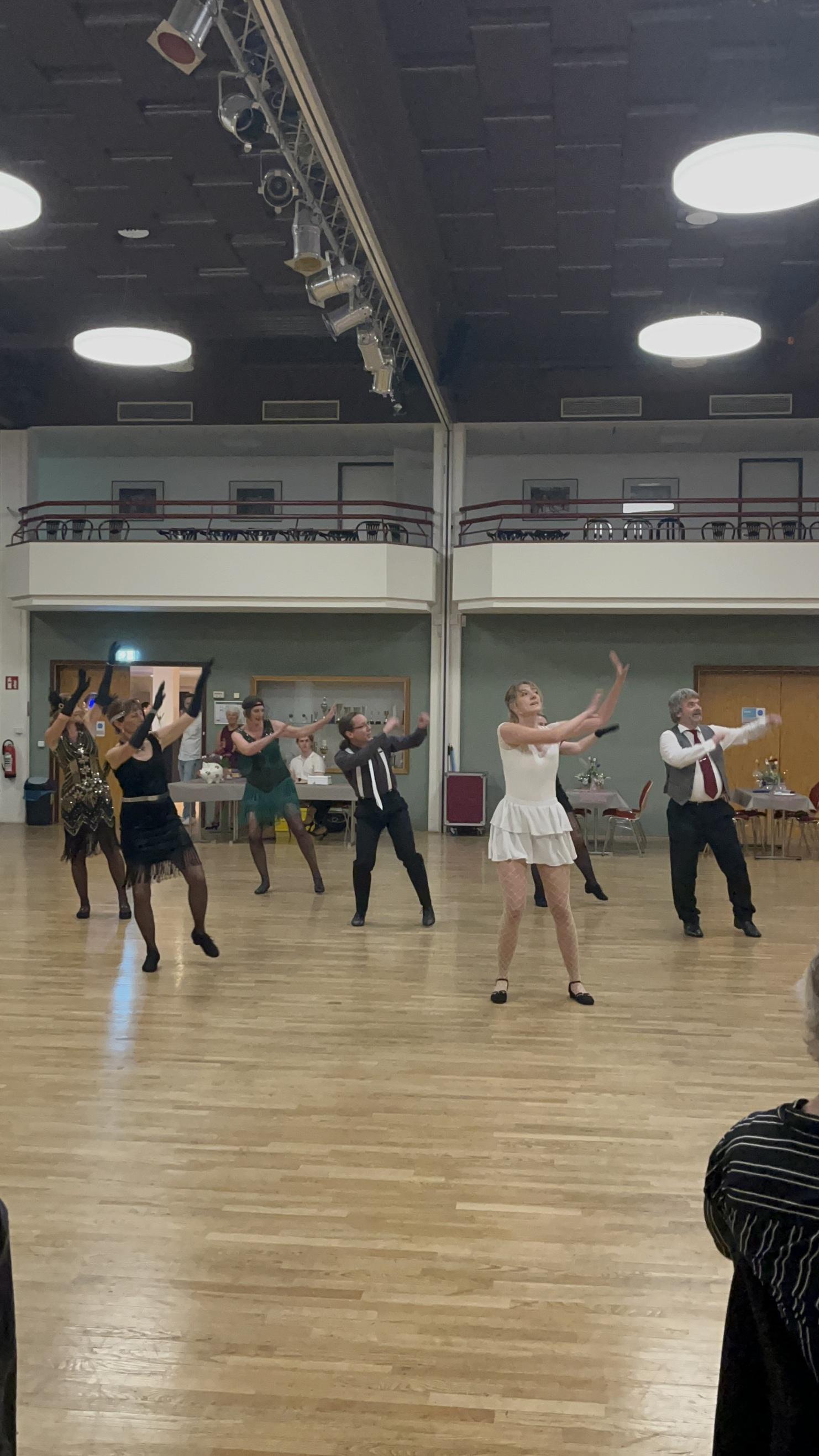 Tanzgruppe in eleganter Kleidung führt eine Choreografie auf einer Tanzfläche vor einem Publikum auf.