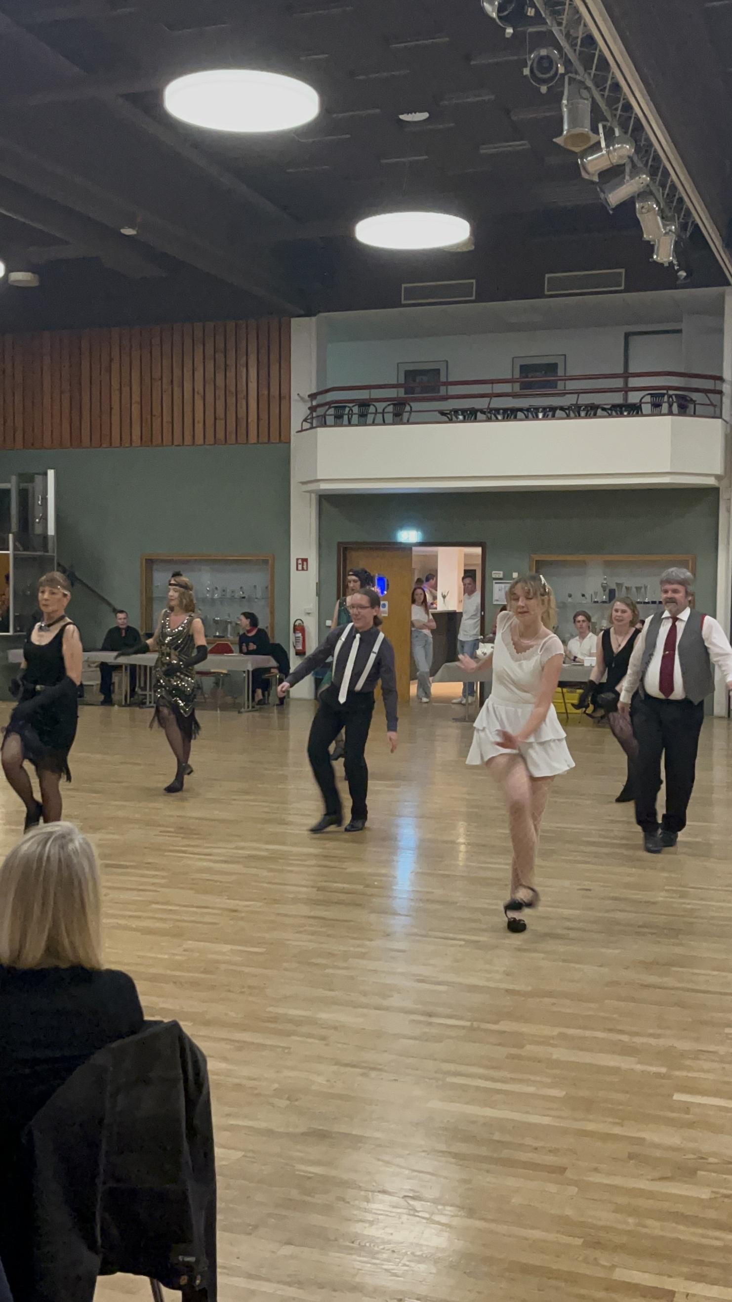 Tanzgruppe in eleganten Kleidern bewegt sich auf einem Holzfußboden während einer Veranstaltung in einem Saal.