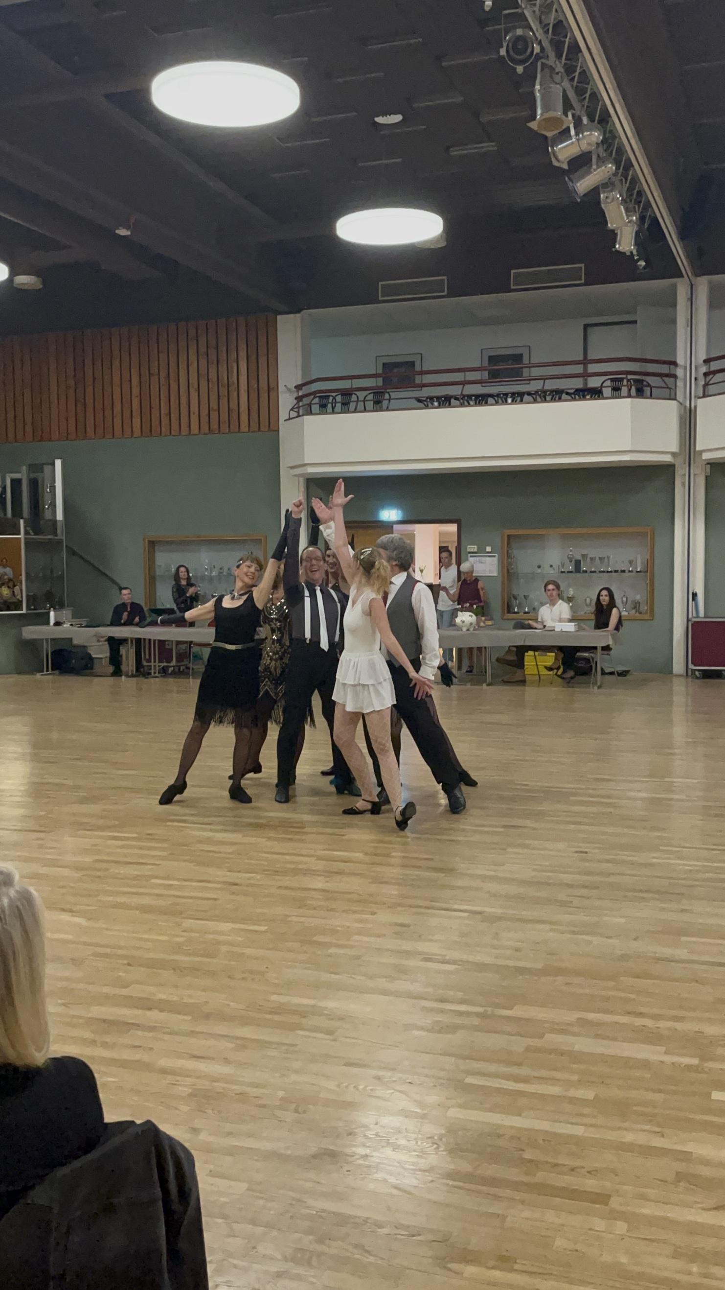 Tanzgruppe in eleganter Kleidung, die auf einer Holzfläche eine tänzerische Aufführung präsentiert.