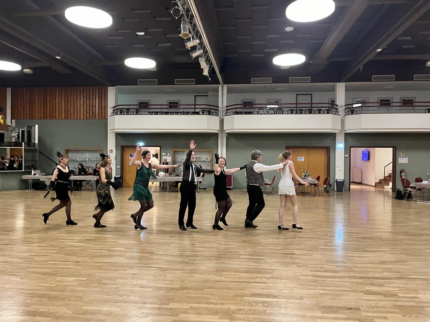 Tanzgruppe in festlicher Kleidung führt eine Choreografie auf einer holzlatten Tanzfläche aus.