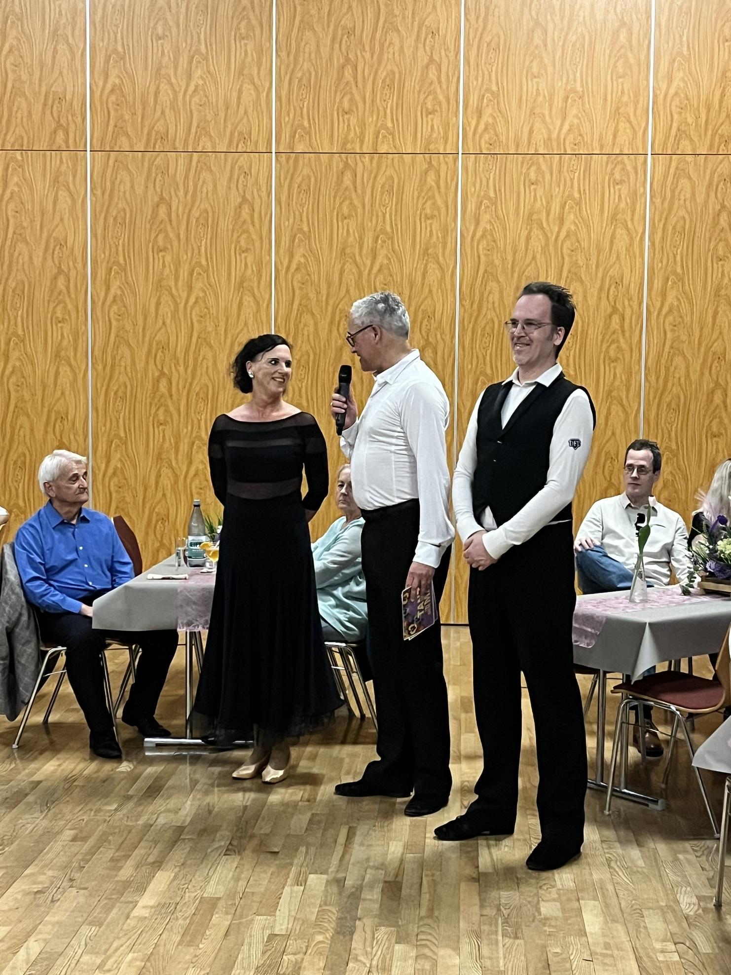 Paar steht auf einer Veranstaltung, während ein Mann mit Mikrofon spricht. Gäste sitzen an Tischen im Hintergrund.