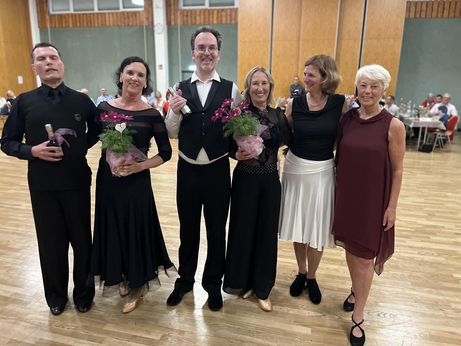 Fünf Personen in eleganter Kleidung posieren lächelnd, halten Blumensträuße und eine Flasche Sekt in einem festlichen Saal.