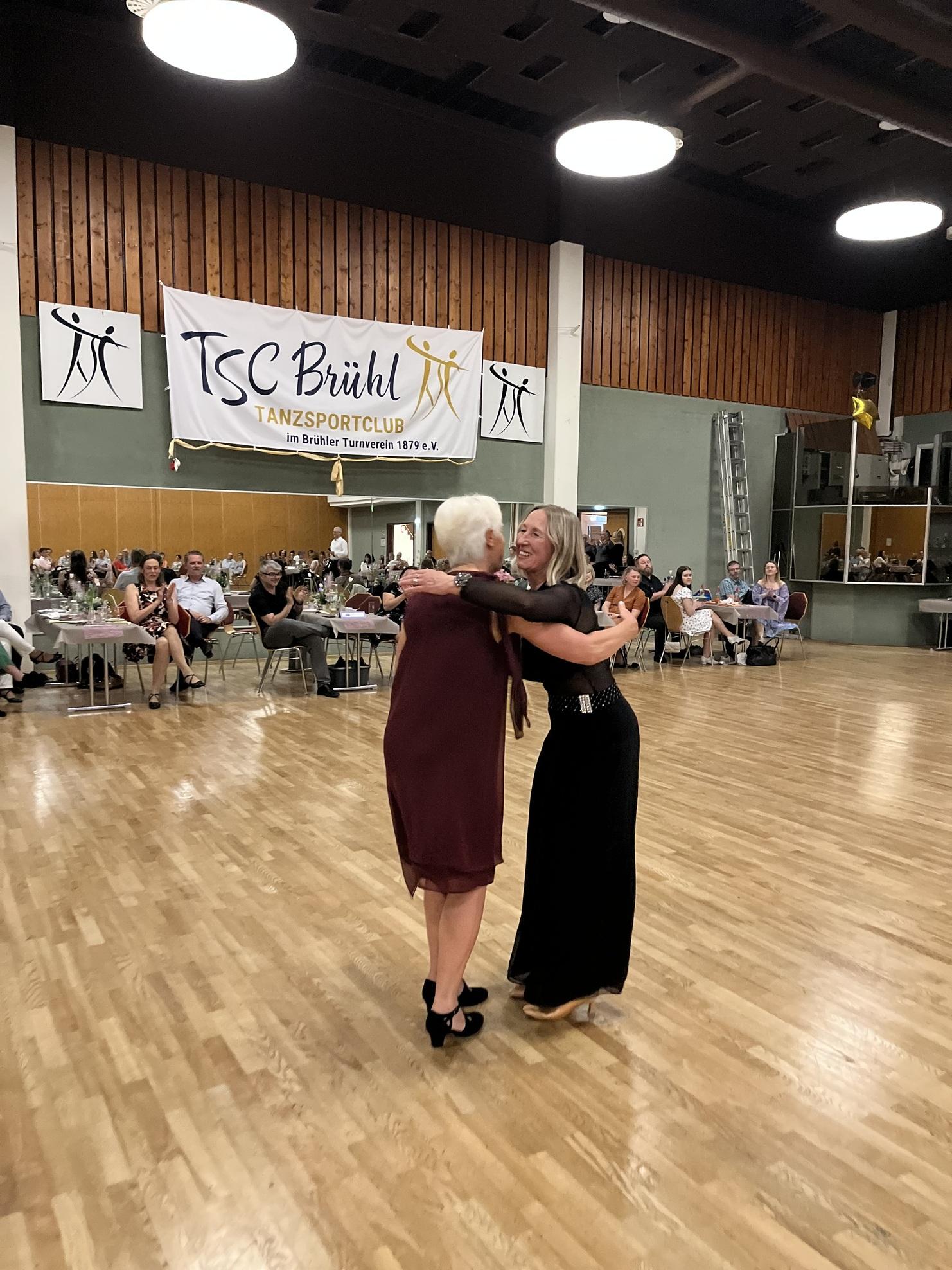 Zwei Frauen umarmen sich in einem Festsaal des Tanzsportclubs Brühl, im Hintergrund tanzende Gäste und Dekoration.