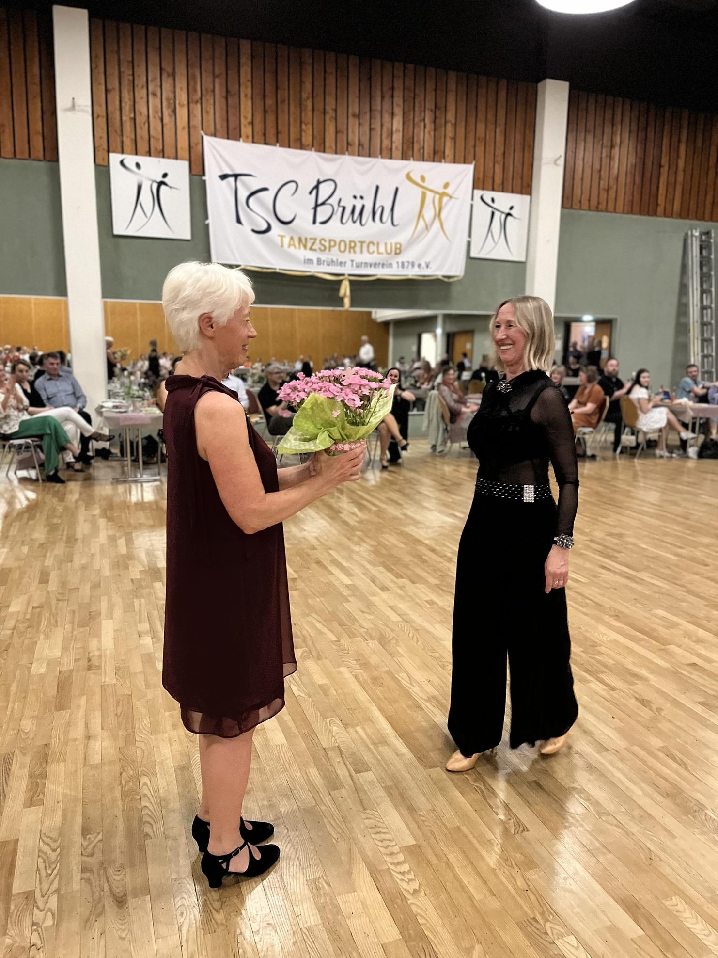 Zwei Frauen stehen sich lächelnd gegenüber, eine überreicht der anderen einen Blumenstrauß, im Hintergrund ein festlich dekorierter Raum.