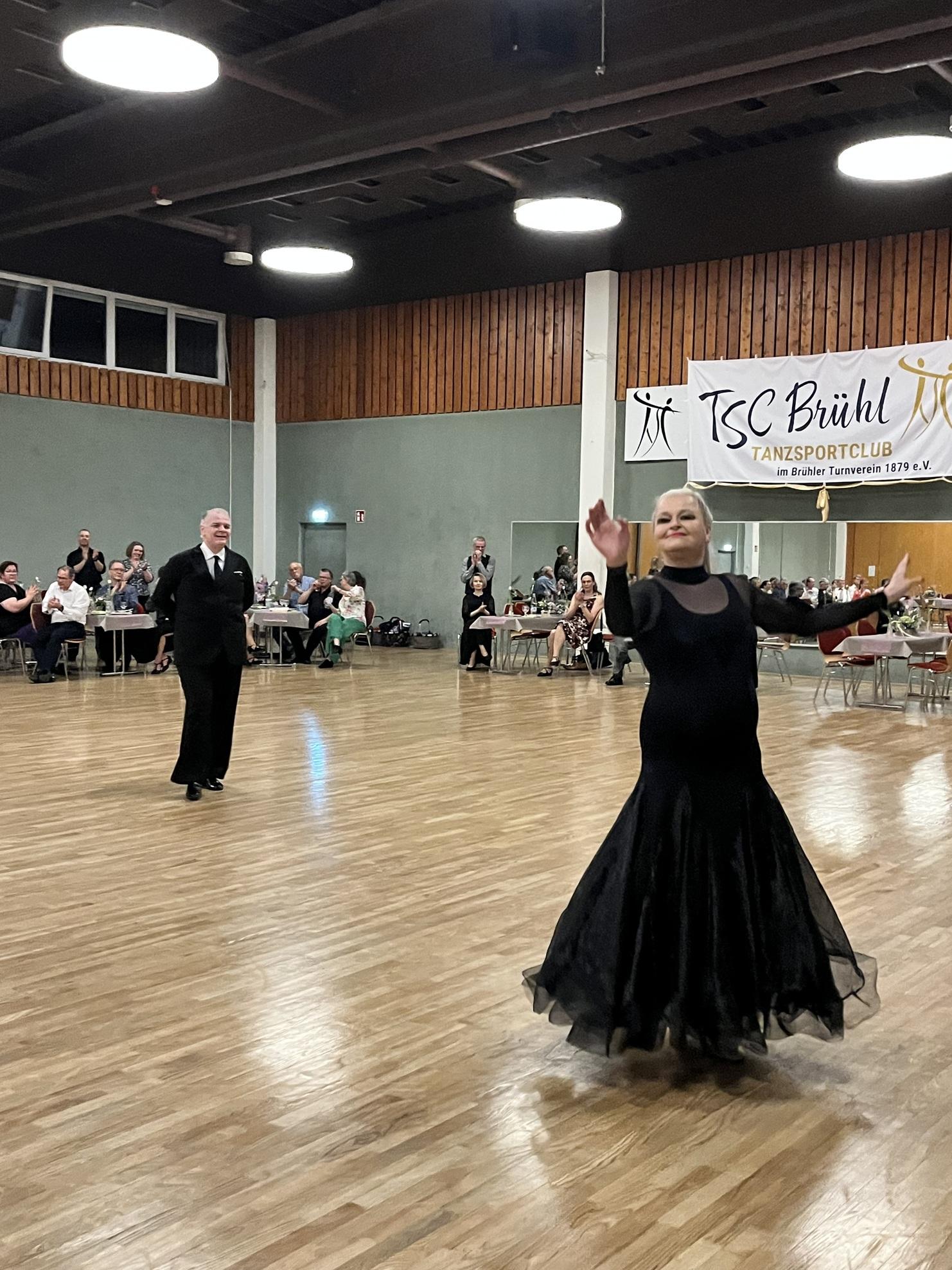 Tanzpaar in eleganter Kleidung führt einen Tanz in einer festlichen Atmosphäre mit Zuschauern im Hintergrund auf.