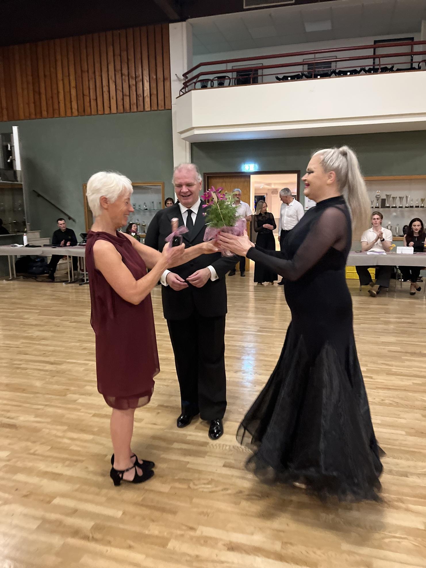 Zwei Frauen überreichen ein Geschenk mit Blumen, während ein Mann im Hintergrund lächelt. Tanzveranstaltung in einem Saal.