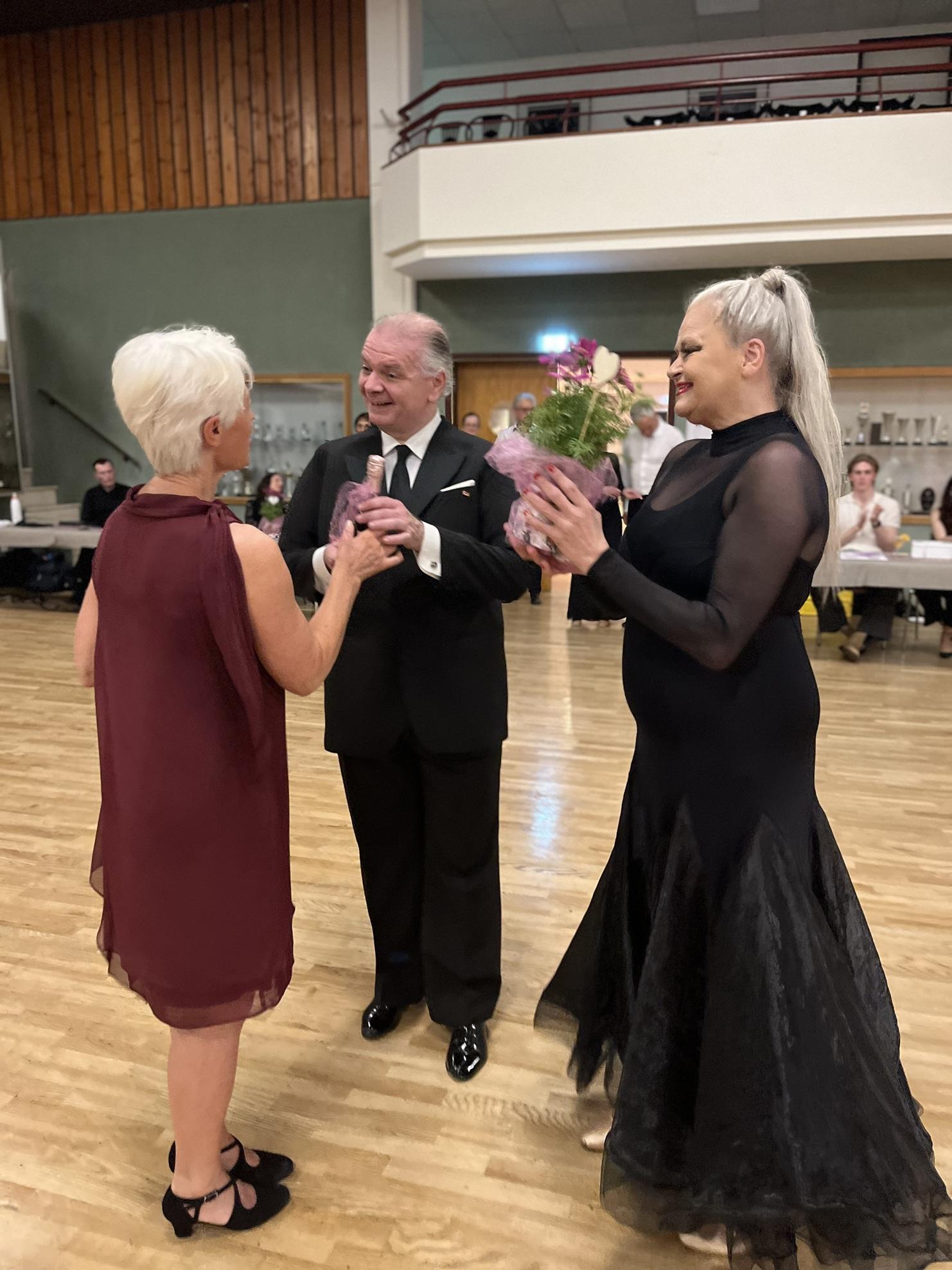 Drei Personen stehen in festlicher Kleidung, eine überreicht Blumen, im Hintergrund ist eine tanzende Menge sichtbar.