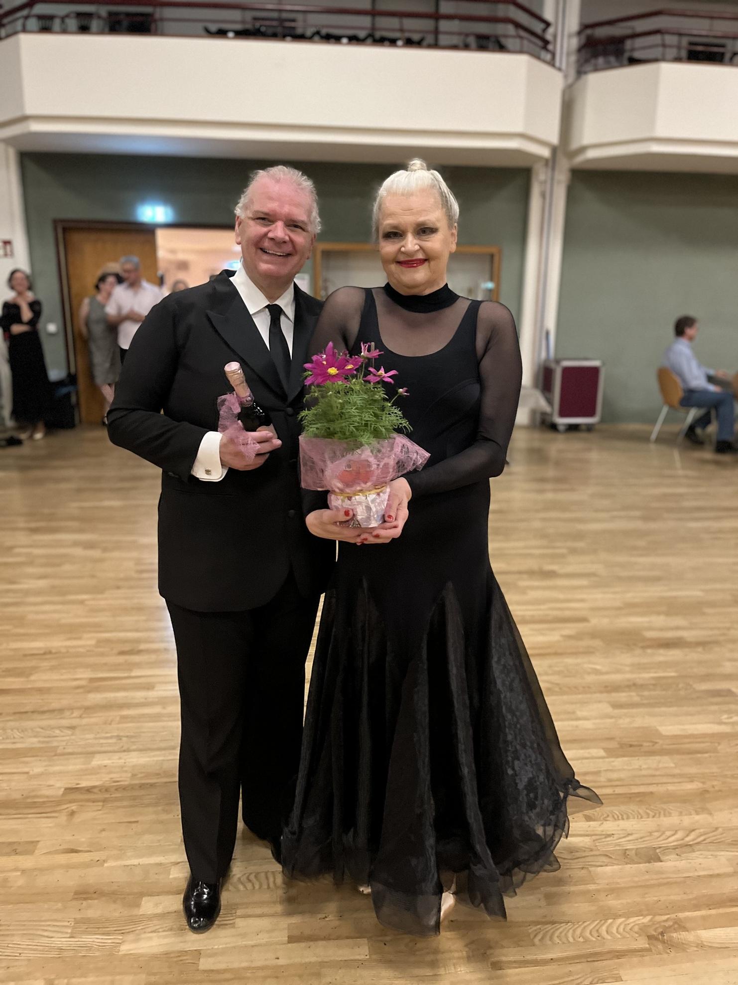 Paar in eleganter Kleidung posiert lächelnd, der Mann hält eine Flasche, die Frau einen Blumenstrauß in Geschenkpapier.