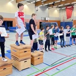 Kinder stehen in einer Turnhalle auf Podesten und halten Urkunden in der Hand, während sie auf eine Veranstaltung warten.