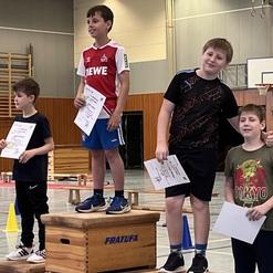 Vier Kinder stehen auf einem Siegerpodest in einer Sporthalle, halten Urkunden in der Hand und lächeln in die Kamera.