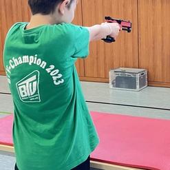 Junge in grünem T-Shirt zielt mit einer Sportpistole auf eine Zielscheibe in einer Turnhalle.