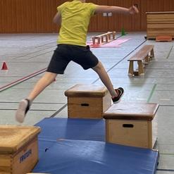 Junge springt über zwei Turnkästen in einer Sporthalle auf blauen Matten. Bunte Hütchen sind im Hintergrund platziert.