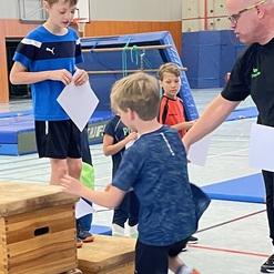 Kinder trainieren in einer Turnhalle, ein Junge klettert auf eine Holzsprungbank, während ein Trainer Anweisungen gibt.