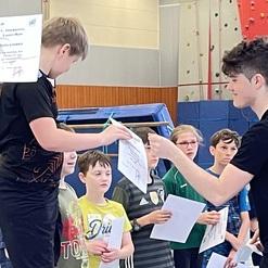 Ein Junge erhält eine Urkunde von einem Erwachsenen in einer Turnhalle, umgeben von weiteren Kindern.