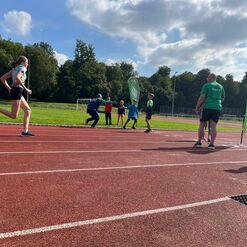 Joggerin sprintet auf der Laufbahn während Trainer und Kinder an der Seite stehen, mit Wettkampf-Bannern im Hintergrund.