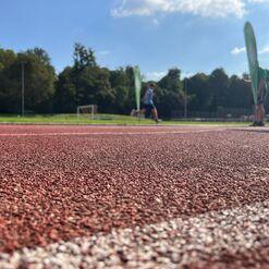 Nahaufnahme der roten Laufbahn mit einem Athleten, der im Hintergrund läuft, und einer Person mit Grünem Banner.