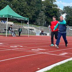 Zwei Männer laufen fröhlich auf einer roten Laufbahn, ein Zelt und Zuschauer im Hintergrund.