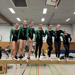 Sechs Turnerinnen stehen auf einem Barren, tragen einheitliche schwarze Anzüge mit grünen Ärmeln und lächeln in die Kamera.