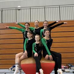 Sechs Turnerinnen in sportlicher Kleidung posieren auf einem Gerät im Gymnastikraum, mit Holzpaneelen im Hintergrund.