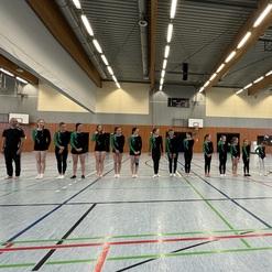 Gruppe von Gymnastinnen in Sportkleidung steht aufgereiht in einer Sporthalle neben einem Trainer und Turngeräten.