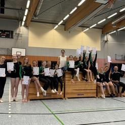 Gruppe von Turnerinnen in Sportbekleidung hält Urkunden in einer Turnhalle, mit einem Trainer auf einer Turnbank.