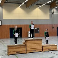 Drei Gymnastinnen stehen auf Podesten, halten Urkunden und tragen enge, schwarz-grüne Sportanzüge in einer Sporthalle.