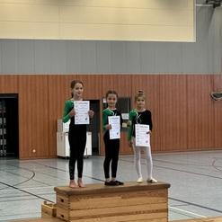 Drei Mädchen in grünen Gymnastikanzügen halten Urkunden auf einem Holzpodest in einer Sporthalle.