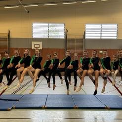 Gruppe von Turnerinnen in Trainingsanzügen, die auf einem Schwebebalken sitzen, lächeln gemeinsam in einer Turnhalle.