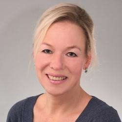 Porträt einer lächelnden Frau mit blonden Haaren, die einen blauen Pullover trägt, vor neutralem Hintergrund.