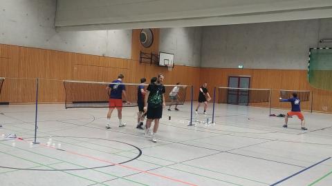 Sechs Personen spielen Badminton in einer Sporthalle mit zwei Netzten und Sicht auf Basketballkörbe.